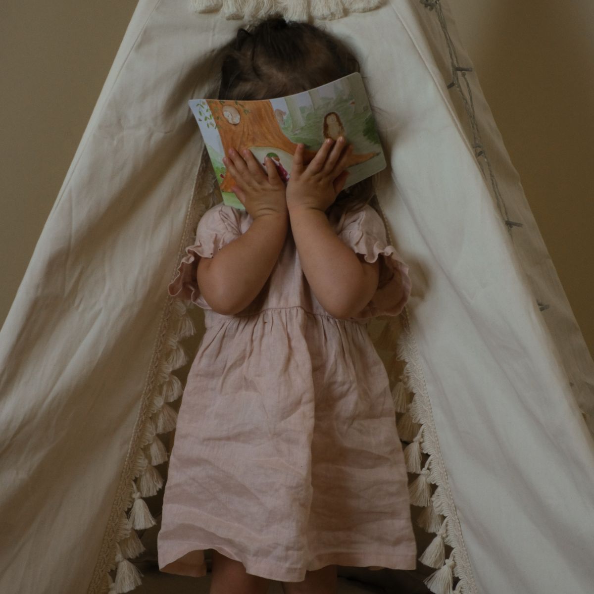Personalised snail mail for children from Tilly’s Letters. A toddler in a light pink dress sits inside a cream fabric teepee tent, holding a children's book over their face.