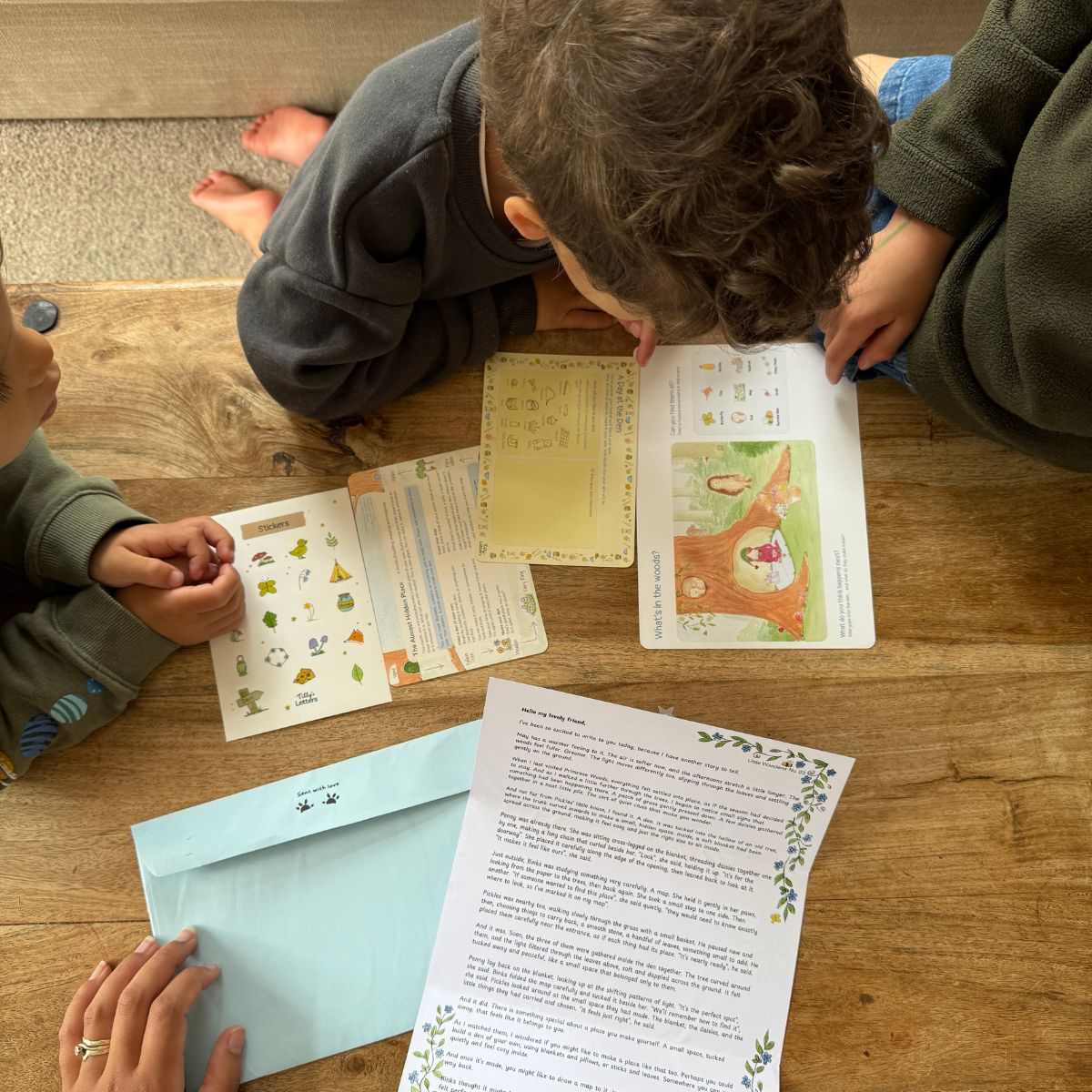 Personalised envelope addressed to a child by name. Children gathered on a wooden floor around printed activity sheets, a light blue envelope, and a letter, suggesting a homeschooling or educational craft project.