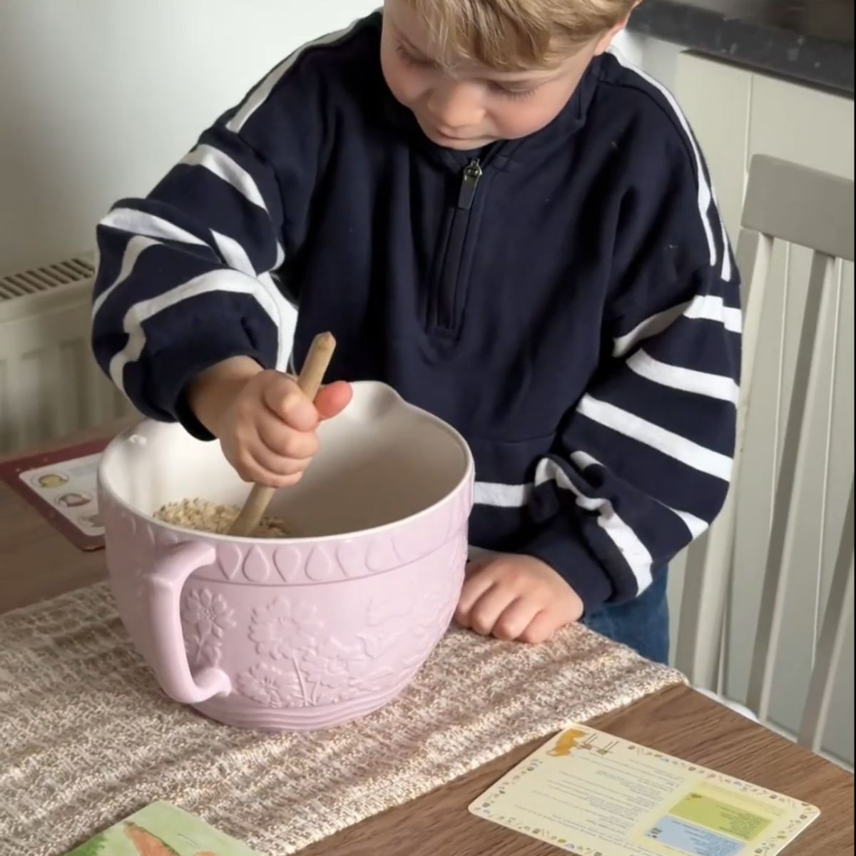 Screen-free snail mail for children with stickers and story card. A young boy with blond hair wearing a navy blue and white striped quarter-zip sweatshirt is standing at a table, mixing ingredients in a large light pink ceramic bowl with a wooden spoon.