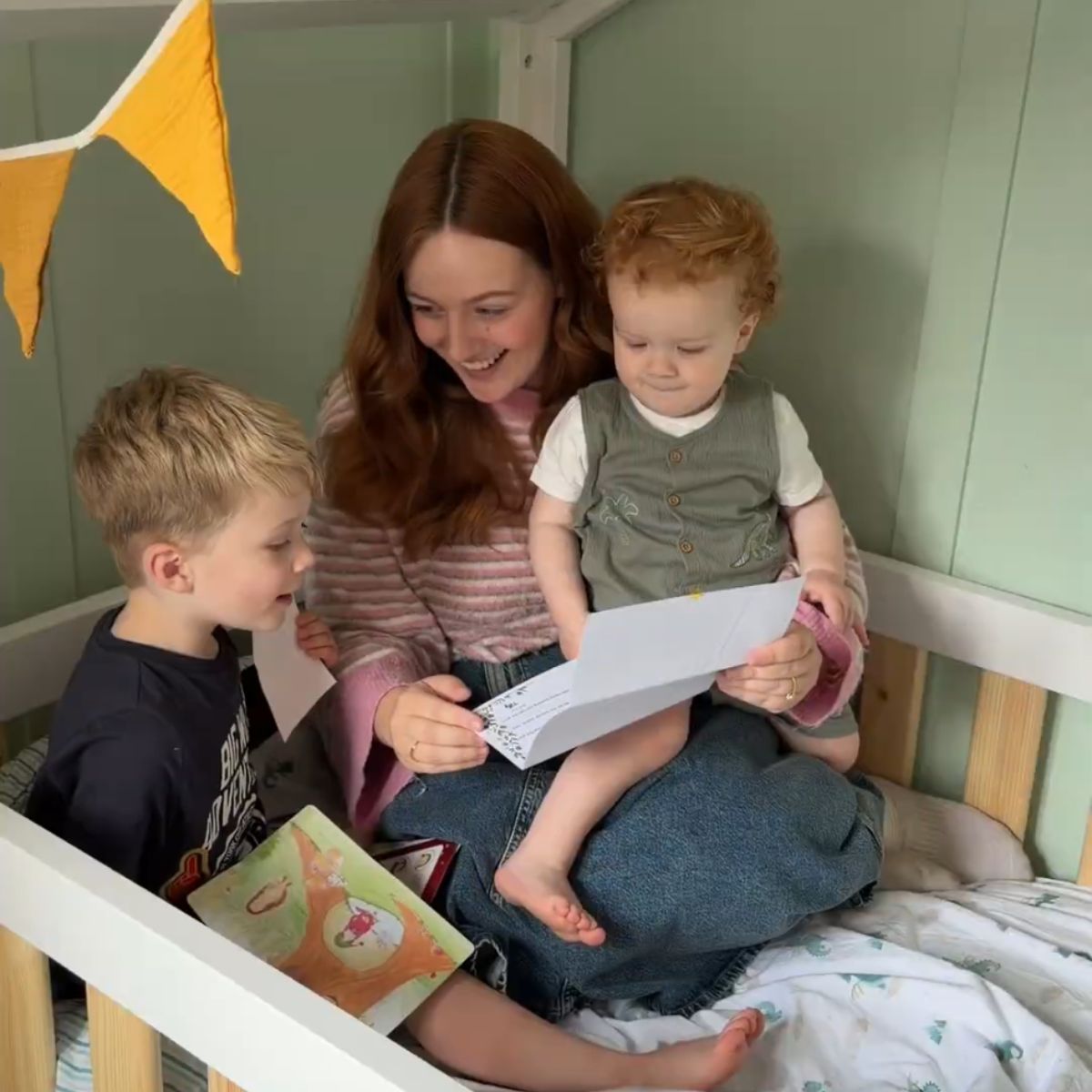 Monthly letter subscription for children aged 2 to 6. A woman sits in a wooden bed frame reading a sheet of paper to two young children, one boy and one girl, in a cozy bedroom setting with a green paneled wall and yellow bunting.
