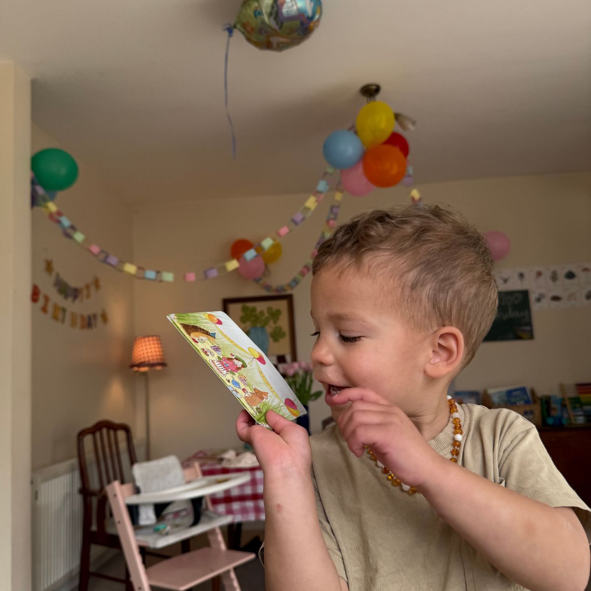 Letterbox gift for children with story, recipe and activity cards. A young child in a beige t-shirt and amber bead necklace holds up a colourful illustrated card in a decorated birthday room with balloons and party banners.